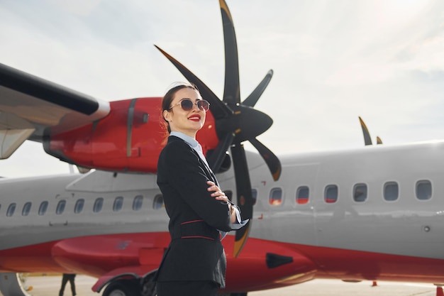 Young stewardess that is in formal black clothes is standing outdoors near plane