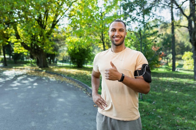 Young strong hispanic man standing in headphones and on phone outdoors and smiling pointing finger