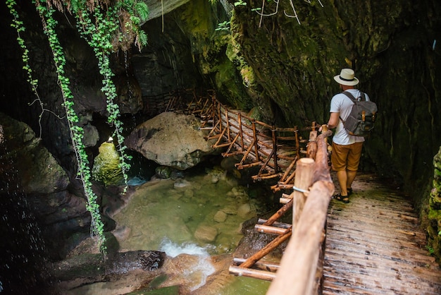 young strong man in white hat with backpack at cave entrance hiking concept creek with waterfall