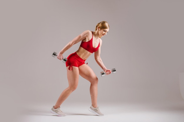 Young strong and sporty woman in red cloth doing exercises with dumbbells on gray background