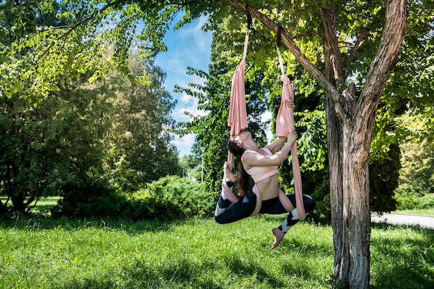 Young strong woman with a thin waist with strong arms engaged in exercises on an air hammock in nature Outdoor fly yoga concept