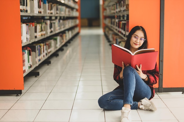 young student in casual style sitting on the floor by bookshelf while holding a book in the library