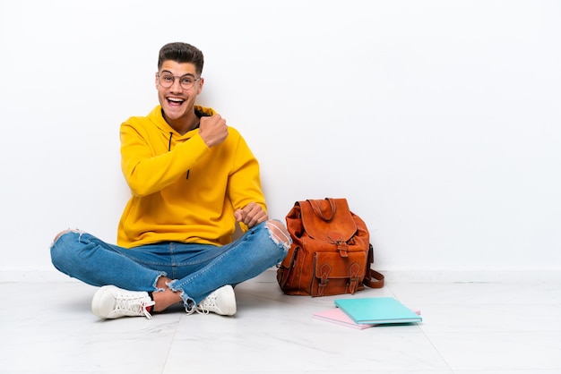 Young student caucasian man sitting one the floor isolated on white background celebrating a victory