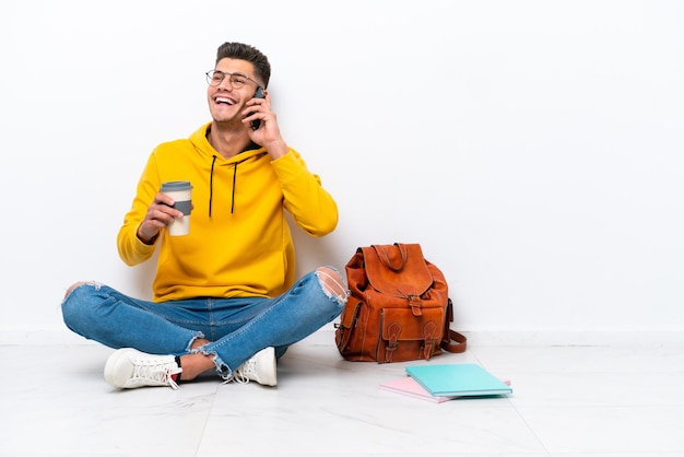 Young student caucasian man sitting one the floor isolated on white background holding coffee to take away and a mobile