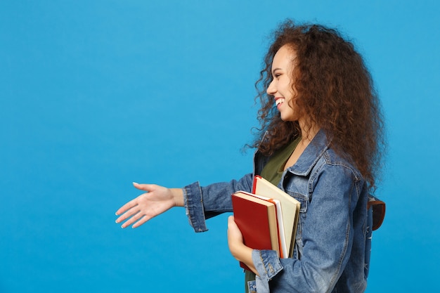 Young student in denim clothes and backpack holds books isolated on blue wall