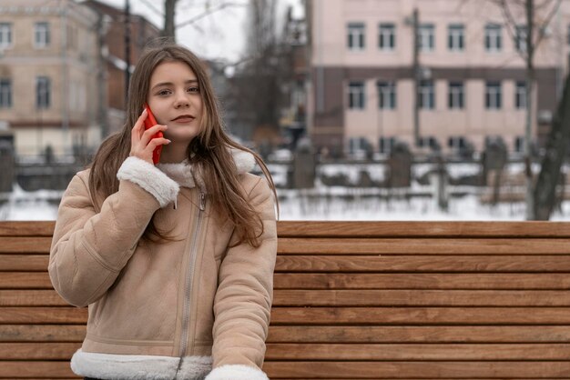 Young student girl sits on park bench in city and talks on phone Woman in beige sheepskin coat rests outside in winter