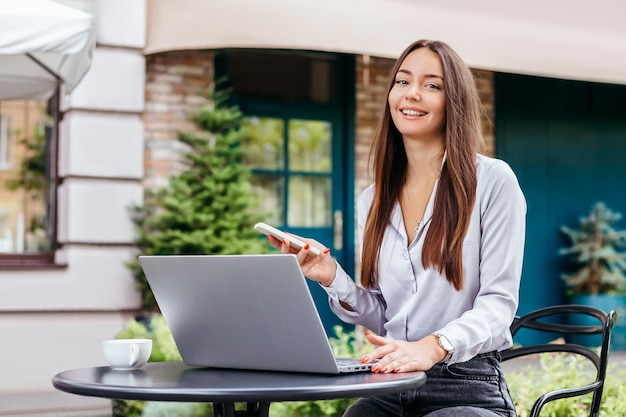 A young student girl uses laptop and a smartphone and looks at the camera