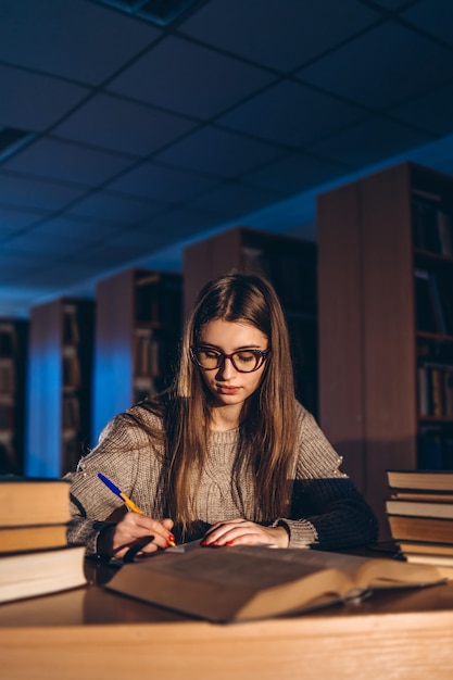 Young student in glasses preparing for the exam. Girl in the evening sits at a table in the library with a pile of books