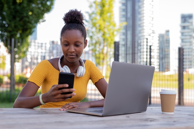 Young student studying in the open air sitting on a table using smartphone and laptop multiple connections freelance black woman connected to the internet