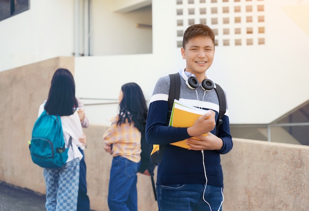 Young student teenager boy or high school student carrying schoolbag holding notebooks with friends in background