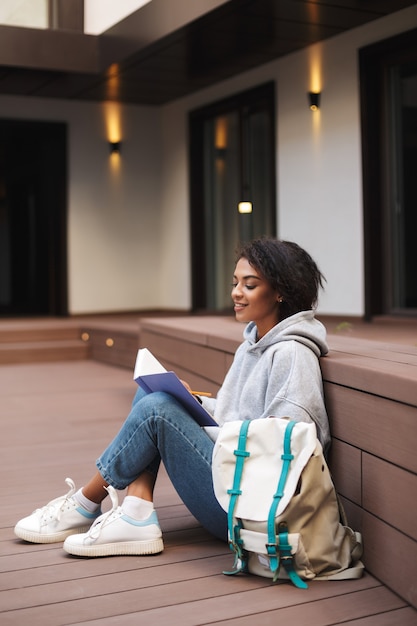 Young student with dark curly hair sitting on floor and writing in notebook. Pretty girl studying alone in courtyard of university