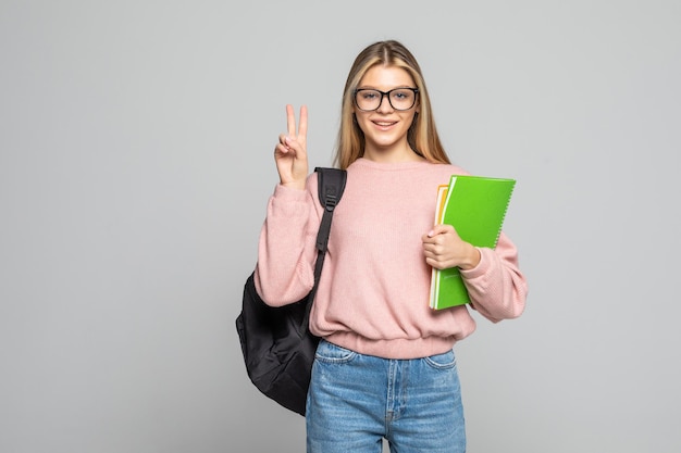 Young student woman doing peace sign with fingers, excellent symbol wearing backpack holding books over isolated on white wall