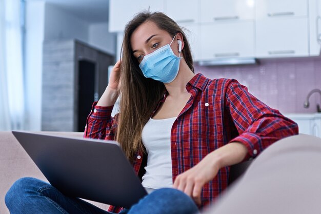 Young student woman in a medical protective mask surfing and watching video online at home at the computer during self isolation and quarantine.