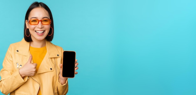 Young stylish chinese woman showing mobile phone screen and thumbs up smartphone app standing over blue background