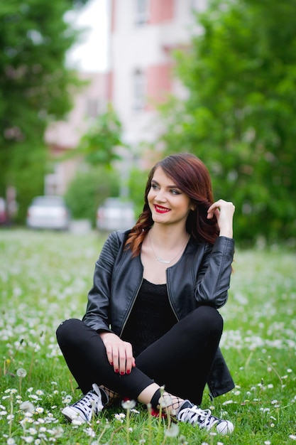 Young stylish female wearing jeans with a leather jacket and sitting on the ground in a park