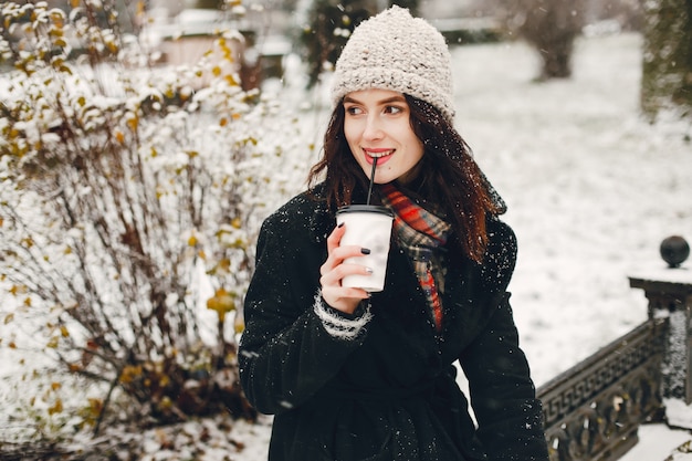 young and stylish girl in black coat and white hat drinking coffee in winter city