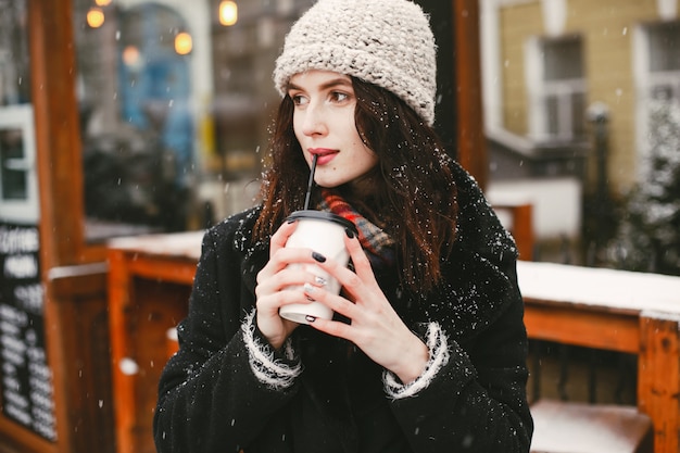 young and stylish girl in black coat and white hat drinking coffee in winter city