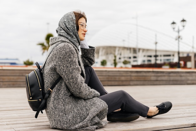 Young stylish girl in gray coat and glasses sits outdoors, cloudy weather, street style