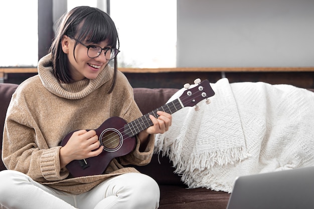 Young stylish girl with glasses learns to play the ukulele. Concept of online education, home education.