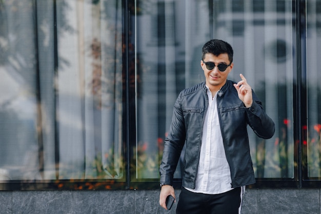 Young stylish guy in glasses in black leather jacket with phone on glass 