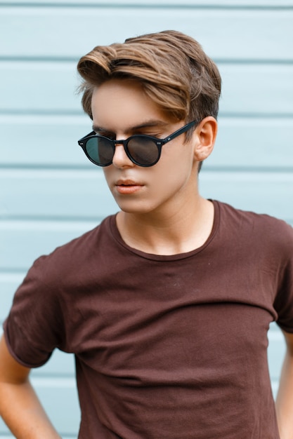 Young stylish handsome hipster man with trendy hairstyle in fashionable brown t-shirt in black sunglasses is posing near a vintage wooden blue wall