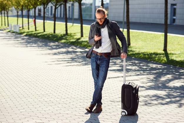 Young stylish man using his smartphone while standing with suitcase near exit the airport outdoors. Business style, traveler, modern lifestyle, business trip.