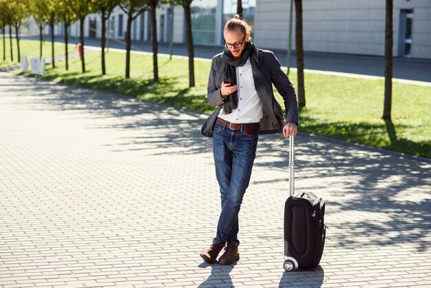 Photo young stylish man using his smartphone while standing with suitcase near exit the airport outdoors. business style, traveler, modern lifestyle, business trip.