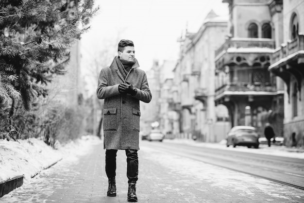 Young stylish man in warm gray coat and leather gloves walking down the street. Street style. Street style. Black and white picture.