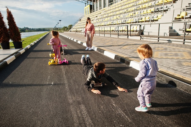 Young stylish mother with four kids outdoor. Sports family spend free time outdoors with scooters and skates.