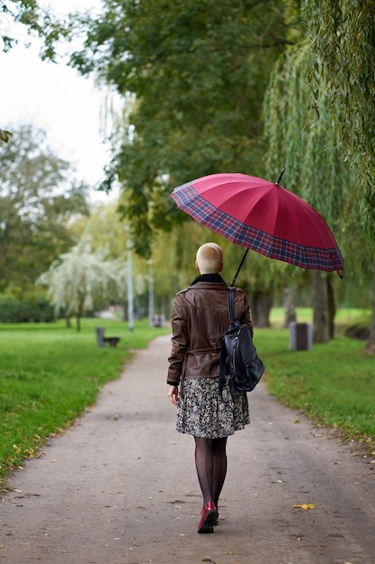 Young stylish short haired blonde woman walks in the autumn park with red umbrella View from back