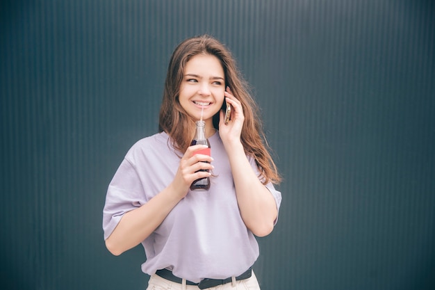 Young stylish trendy woman isolated over grey blue background. Girl talking on phone and drinking soda dark sparkling water from glass bottle through plastic straw.