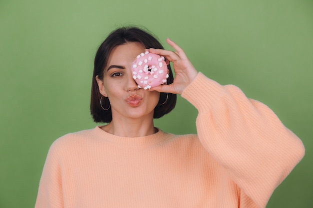 Young stylish woman in casual peach sweater  isolated on green olive wall send kiss with pink donut copy space