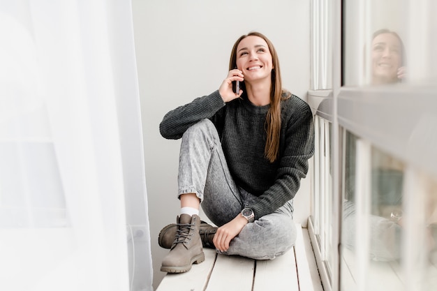 Young stylish woman sitting near window talking on the phone and smiling