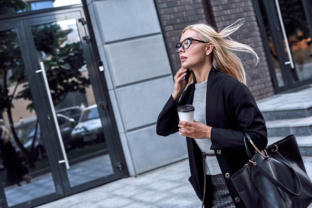 Young stylish woman talking by smartphone she walks with morning coffee