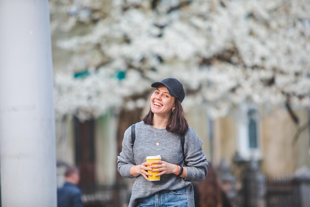 Young stylish woman walking by street with coffee cup spring blooming tree on background