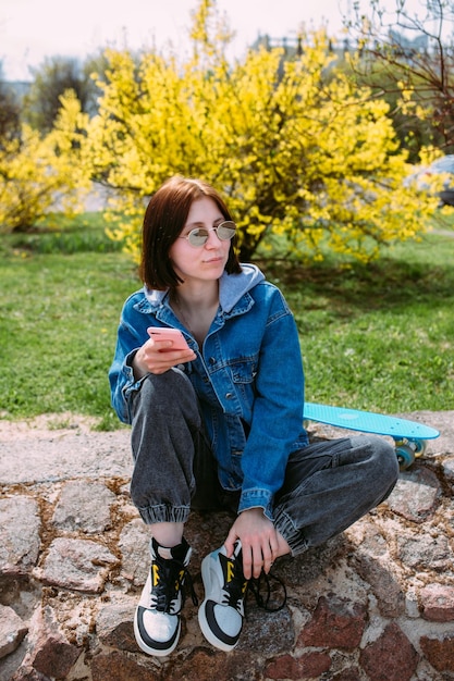 Young stylish woman walks on street with a skateboard and uses a smartphone