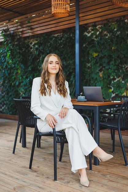 A young stylish woman in a white classic suit with a smile on her face is standing on the terrace of a summer cafe