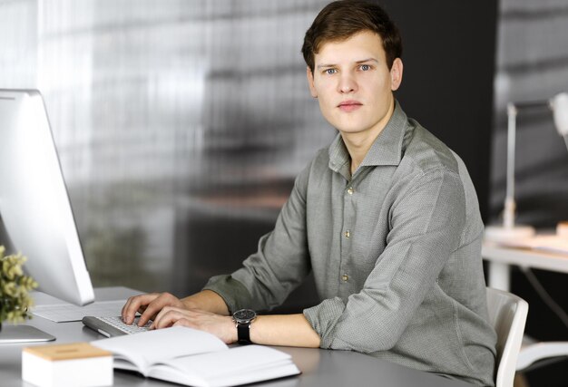 Young successful businessman and programmer in a green shirt is looking at camera, while he is working on his computer, sitting at the desk in a cabinet. Headshot or business portrait in an office.