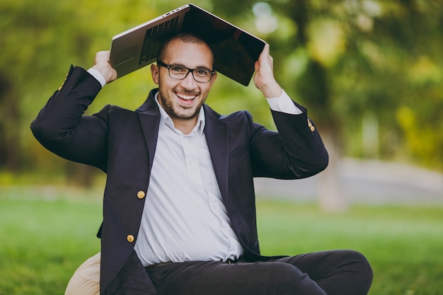 Young successful businessman in white shirt, classic suit, glasses. Man sit on soft pouf under cover laptop pc computer in city park on green lawn outdoors on nature. Mobile Office, business concept.