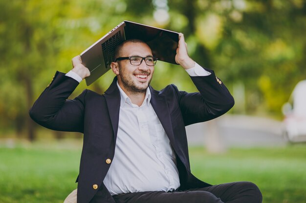 Young successful businessman in white shirt, classic suit, glasses. Man sit on soft pouf under cover laptop pc computer in city park on green lawn outdoors on nature. Mobile Office, business concept.