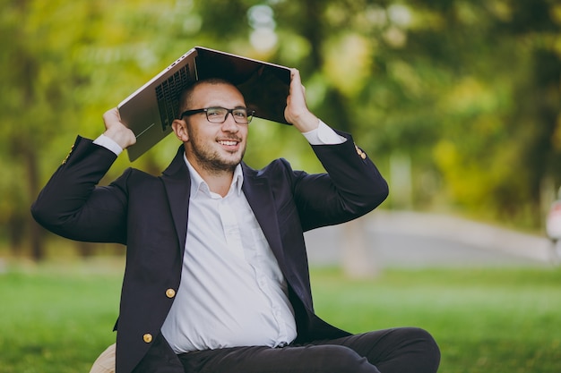 Young successful businessman in white shirt, classic suit, glasses. Man sit on soft pouf under cover laptop pc computer in city park on green lawn outdoors on nature. Mobile Office, business concept.