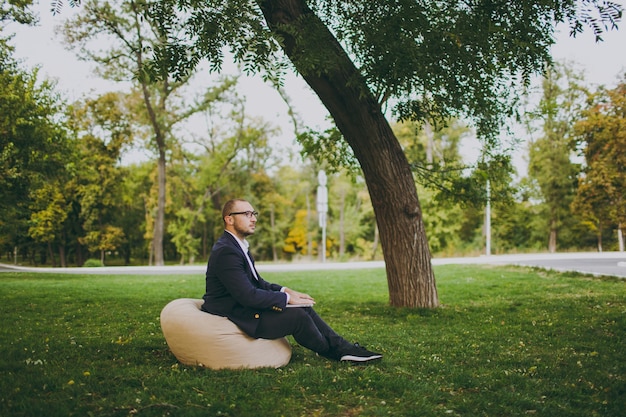 Photo young successful businessman in white shirt, classic suit, glasses. man sit on soft pouf, working on laptop pc computer in city park on green lawn outdoors on nature. mobile office, business concept.