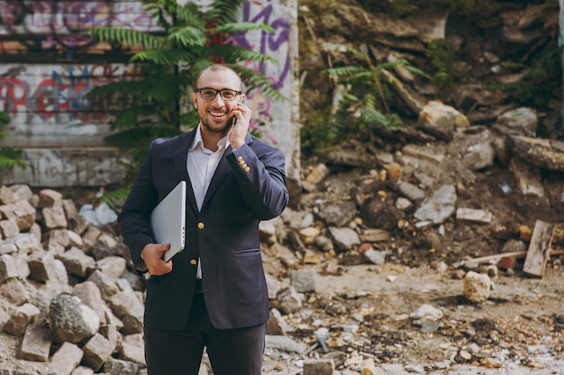 Young successful businessman in white shirt, classic suit, glasses. Man standing with laptop pc computer, talking on phone near ruins, debris, stone building outdoors. Mobile Office, business concept.