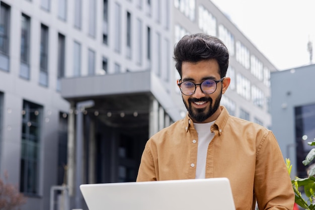 Young successful businessman working with laptop remotely sitting on bench outside office building