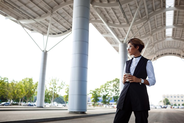 Young successful businesswoman in glasses holding coffee, standing near business centre.