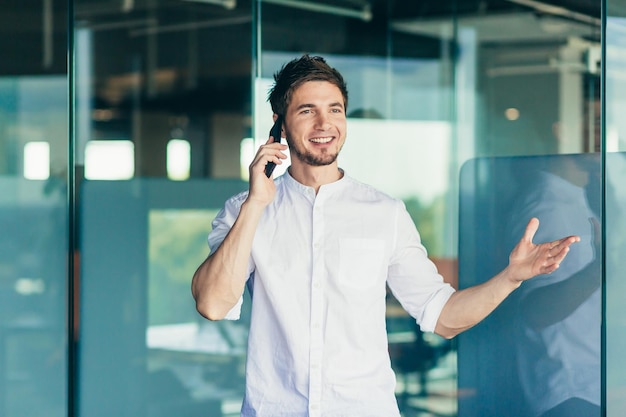 Young successful businesswoman talking by the window in the office on the phone man smiling and wearing a shirt