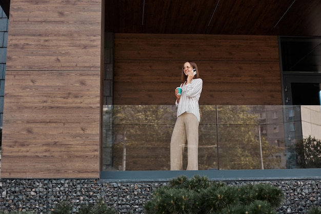 Young successful businesswoman wearing stylish clothing standing near building talking on mobile phone smiling girl making business call on cell