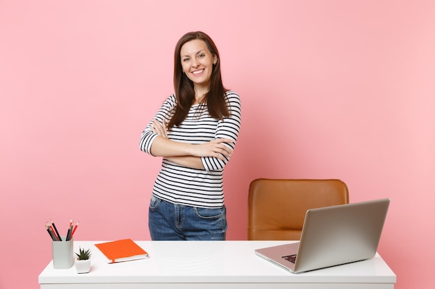 Young successful girl in casual clothes work standing near white desk with contemporary pc laptop isolated on pastel pink background. Achievement business career concept. Copy space for advertisement.