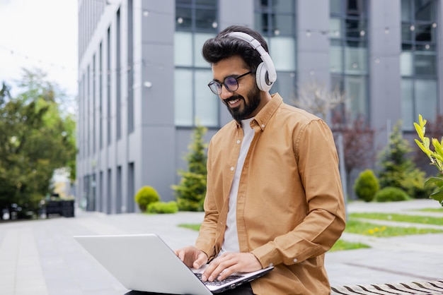 Young successful hindu student studying online using laptop man wearing headphones listening to