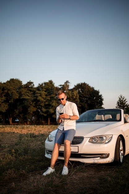 Young successful male businessman is standing near his white convertible car. A guy in sunglasses in a white shirt and shorts near his car.